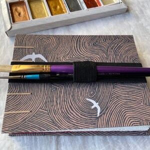 May include: A watercolor painting set with a sketchbook and paintbrushes. The sketchbook has a brown and gray patterned cover with white bird illustrations. The paintbrushes are held together with a black elastic band. A palette of metallic watercolors is visible.