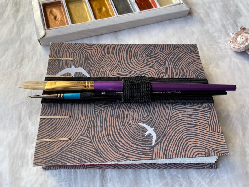 May include: A watercolor painting set with a sketchbook and paintbrushes. The sketchbook has a brown and gray patterned cover with white bird illustrations. The paintbrushes are held together with a black elastic band. A palette of metallic watercolors is visible.