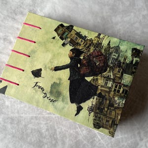 Könnte beinhalten: Ein Notizbuch mit einer skurrilen Illustration einer Person mit Rucksack, die über einer Stadtlandschaft schwebt. Der Einband ist hellgrün mit rosa Nähten und dem Wort "Imagine" in Schreibschrift.