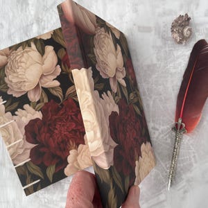 Pode incluir: Um diário floral com um design de peónias em creme e bordô. Uma pena decorativa e uma concha estão ao lado do diário. O diário está aberto, revelando a encadernação costurada.