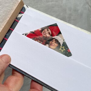 May include: A small, handmade photo album with a white interior and a decorative cover. The album is open, revealing a photograph of two people. The binding is stitched with pink thread. The album is held in a hand.