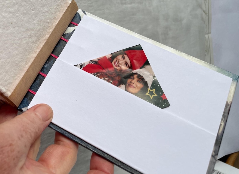 May include: A small, handmade photo album with a white interior and a decorative cover. The album is open, revealing a photograph of two people. The binding is stitched with pink thread. The album is held in a hand.