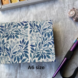 May include: An A6 size notebook with a blue floral pattern. The notebook is bound with metal staples. A purple paintbrush with a black elastic band is next to the notebook. A seashell and a watercolor paint set are also in the image.
