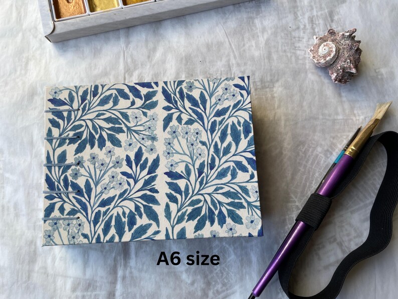May include: An A6 size notebook with a blue floral pattern. The notebook is bound with metal staples. A purple paintbrush with a black elastic band is next to the notebook. A seashell and a watercolor paint set are also in the image.