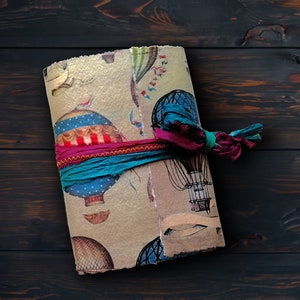 May include: A handmade journal with a vintage hot air balloon design. The cover features a beige background with colorful balloons and flags. A teal and magenta ribbon wraps around the journal, securing it closed. The journal is on a dark wood surface.