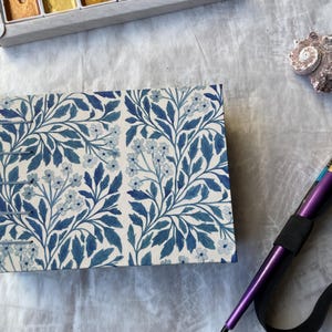 May include: A rectangular notebook with a blue floral pattern, a seashell, and a purple paintbrush with a black elastic band. A watercolor paint set is in the background.