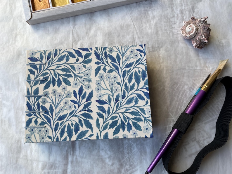 May include: A rectangular notebook with a blue floral pattern, a seashell, and a purple paintbrush with a black elastic band. A watercolor paint set is in the background.