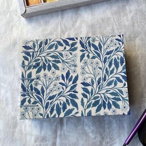 May include: A rectangular journal with a blue floral pattern. The cover features a repeating design of blue leaves and small white flowers on a cream background. The journal is bound with silver metal rings.