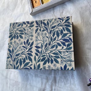May include: A rectangular journal with a floral pattern in shades of blue and white. The design features stylized leaves and small flowers. The journal has a stitched binding and is lying on a white surface.