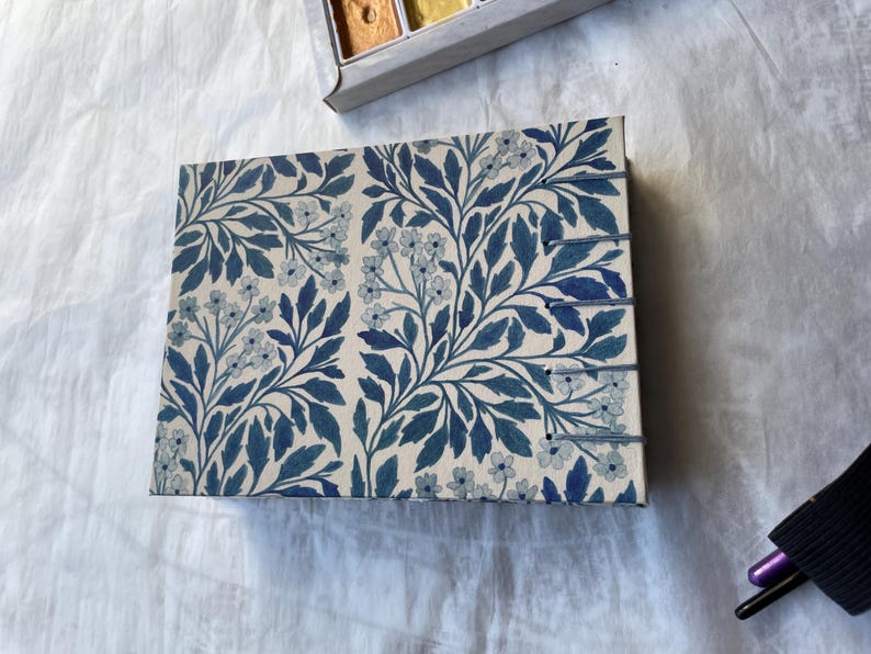 May include: A rectangular journal with a floral pattern in shades of blue and white. The design features stylized leaves and small flowers. The journal has a stitched binding and is lying on a white surface.