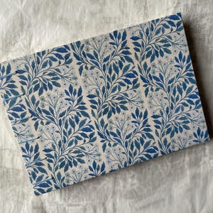 Puede incluir: Un diario rectangular con un estampado floral azul y blanco. La cubierta presenta un diseño repetido de hojas y flores estilizadas en tonos azules sobre un fondo crema. El lomo está cosido a mano con hilo azul.