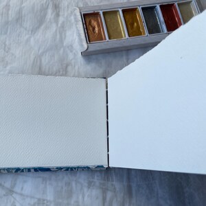 May include: An open sketchbook with textured white pages, next to a palette of metallic watercolor paints. The sketchbook's binding is visible, and the pages are blank, ready for art. The palette contains gold, bronze, and silver hues.