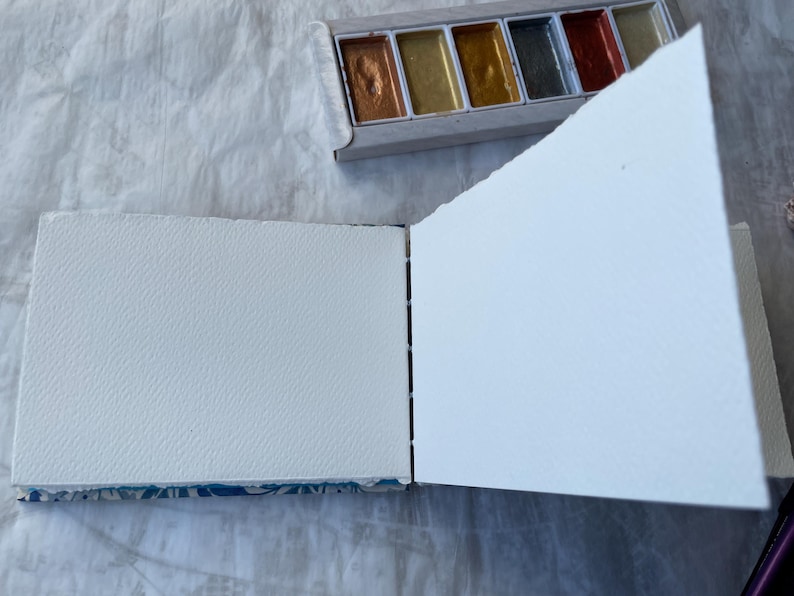 May include: An open sketchbook with textured white pages, next to a palette of metallic watercolor paints. The sketchbook's binding is visible, and the pages are blank, ready for art. The palette contains gold, bronze, and silver hues.