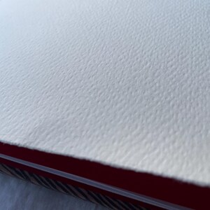 May include: Close-up of a textured, off-white watercolor paper. The paper has a rough, dimpled surface. The edge of the paper is visible, revealing a red and patterned binding.