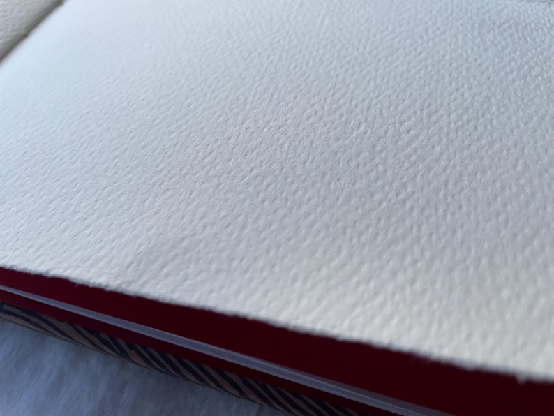 May include: Close-up of a textured, off-white watercolor paper. The paper has a rough, dimpled surface. The edge of the paper is visible, revealing a red and patterned binding.