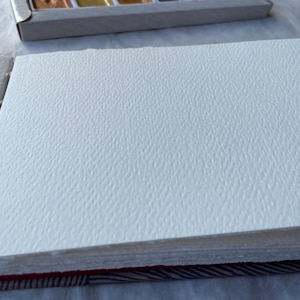 May include: An open sketchbook with thick, textured white paper pages. The pages are bound together with a red and patterned spine. A watercolor paint set is visible in the background.