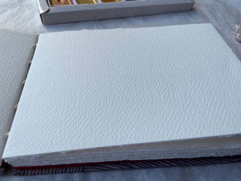 May include: An open sketchbook with thick, textured white paper pages. The pages are bound together with a red and patterned spine. A watercolor paint set is visible in the background.