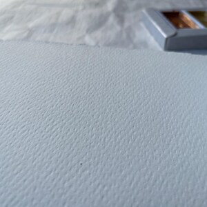 May include: A close-up of a textured, light blue watercolor paper with a deckled edge. A small, silver metal palette with metallic watercolor paints is in the background. The paper has a subtle, dimpled surface.