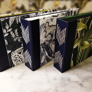 May include: Four hardcover journals with unique covers. The journals feature dark blue spines with white stitching. The covers display various patterns, including floral, marble, and botanical prints. The edges are colored yellow and green.
