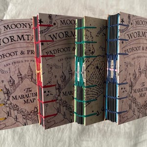 Op de afbeelding: Een stapel handgemaakte journals met bruine covers met tekst en illustraties. De ruggen zijn met de hand gestikt met gekleurd garen in geel, rood, groen en blauw. De tekst bevat "Moony, Wormtail, Padfoot & Prongs" en "The Marauders Map".