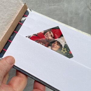May include: Open photo album with a white interior and a photo of two people. The album has a decorative patterned cover and pink stitching along the spine. The photo is visible through a triangular cutout.
