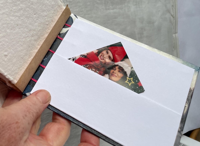 May include: Open photo album with a white interior and a photo of two people. The album has a decorative patterned cover and pink stitching along the spine. The photo is visible through a triangular cutout.