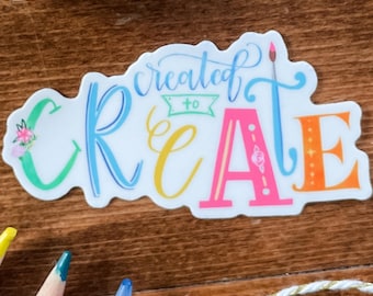 May include: A die-cut sticker featuring the colorful text "created to CREATE" in a mix of blue, green, yellow, pink, and orange lettering. The letter "T" is stylized as a paintbrush, and the "C" includes a small floral accent.