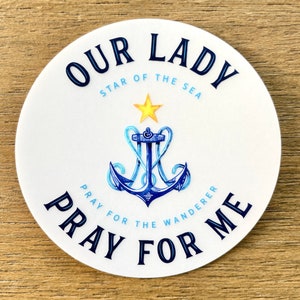 May include: White round sticker with blue and black text that reads "Our Lady Star of the Sea Pray for Me". A blue anchor with a yellow star above it is in the center of the sticker.