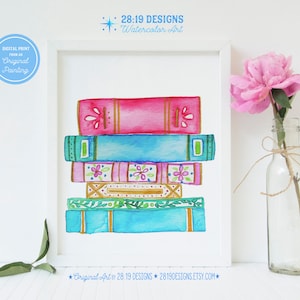 Puede incluir: Impresión artística en acuarela de una pila de libros coloridos en un marco blanco. Los libros están pintados en tonos de rosa, azul y verde con diseños florales y geométricos. Una flor rosa en una botella de vidrio está junto a la obra enmarcada. El texto "28:19 DESIGNS Watercolor Art" está en la parte superior.
