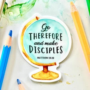 May include: A round sticker featuring a globe design with the text "Go THEREFORE and make DISCIPLES" and "MATTHEW 28:19". The globe has a blue and yellow watercolor design. Blue and yellow pencils are in the background.