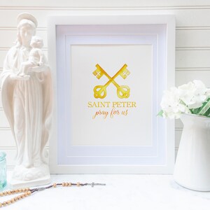 Pode incluir: Uma impressão emoldurada com uma ilustração em aquarela de duas chaves douradas cruzadas, com o texto "SAINT PETER pray for us". A impressão está em uma moldura branca e é exibida com uma estátua branca, um vaso de flores brancas e um rosário.