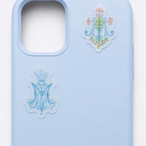 May include: A light blue phone case featuring two decorative stickers. The top sticker displays a blue letter M intertwined with pink lilies. The bottom sticker shows a blue letter M topped with a crown, both set against a scalloped background.