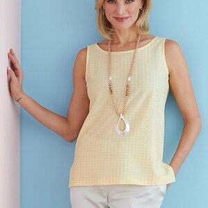 May include: A woman wearing a yellow sleeveless top with a geometric pattern and a long beaded necklace with a silver pendant. She is wearing white pants and has her hair pulled back.