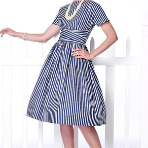 May include: A woman wearing a blue and white striped dress with a white pearl necklace. The dress has a fitted bodice and a full skirt. She is standing in front of a white picket fence.