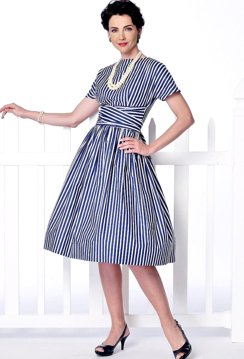 May include: A woman wearing a blue and white striped dress with a white pearl necklace. The dress has a fitted bodice and a full skirt. She is standing in front of a white picket fence.