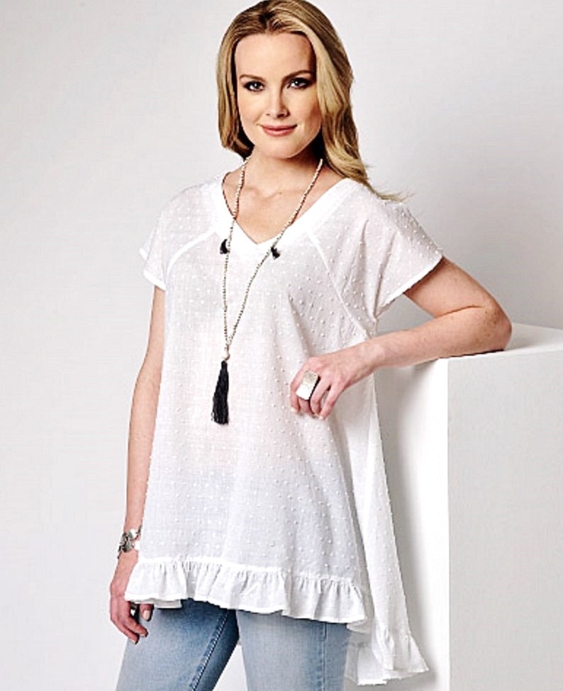 May include: White cotton top with a v-neck, short sleeves, and a ruffled hem. The top has a dotted pattern and a black tassel necklace.