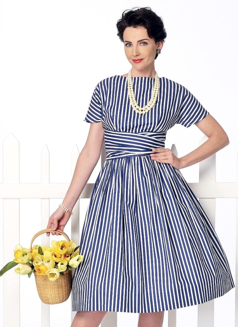 May include: A woman wearing a blue and white striped dress with a white pearl necklace. She is holding a basket of yellow flowers.