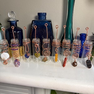 May include: A collection of nine clear glass vials, each wrapped in copper wire that extends upward into a loop. Each vial contains a unique gemstone or crystal, including white, purple, blue, green, yellow, orange, and black stones.