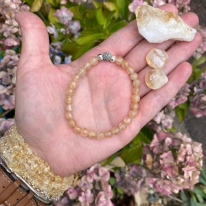 May include: A citrine gemstone bracelet with a silver bead accent. The bracelet is made with small, round beads and is shown on a hand with loose citrine stones.