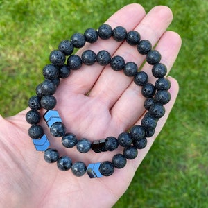 Könnte beinhalten: Zwei schwarze Lava-Armbänder mit silbernen Pfeil-Anhängern. Die Armbänder sind übereinander gelegt.