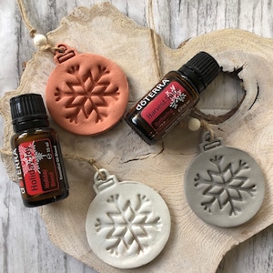 May include: Three ceramic ornaments shaped like Christmas tree baubles with a snowflake design. The ornaments are in shades of red, grey, and white. Two bottles of essential oil are included, one labelled "Holiday Joy" and the other labelled "Harvest Spice".