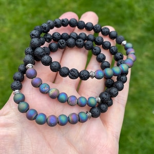 May include: Three bracelets stacked on a hand. The bottom bracelet is made of iridescent agate beads. The top two bracelets are made of black lava rock beads.