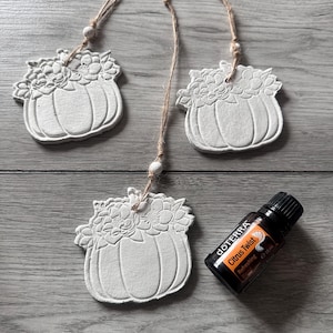 May include: Three white ceramic pumpkin ornaments with floral details hanging from twine. A bottle of doTERRA Citrus Twist essential oil is in the foreground.