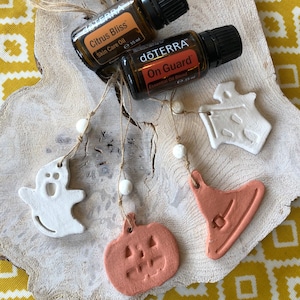 May include: Four Halloween-themed clay ornaments hanging from twine with white beads. The ornaments are a ghost, a witch's hat, a pumpkin, and a house. Two bottles of essential oil are visible in the background. The bottles are labelled "dōTERRA Citrus Bliss Skin Care Oil" and "dōTERRA On Guard Essential Oil Blend".