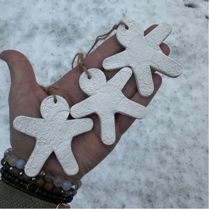 May include: Three white ceramic ornaments shaped like gingerbread people with a raised, textured pattern. The ornaments are hanging from twine.