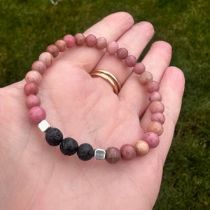 May include: A pink and brown gemstone bracelet with black lava beads and silver spacers. The bracelet is made with round beads and is approximately 7 inches in length.