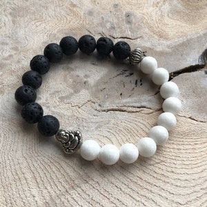 May include: A black and white beaded bracelet with a silver charm in the shape of a seated figure. The bracelet is made with lava rock beads and white beads.