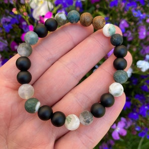 May include: A bracelet made of black, white and green agate beads with a wooden bead. The bracelet is on a person's wrist.