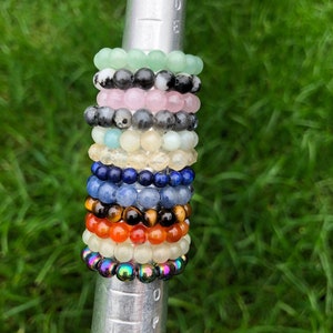 May include: A stack of colourful beaded bracelets, including green, pink, black, blue, and orange. The bracelets are stacked on a metal rod.