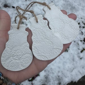 May include: Three white ceramic snowman ornaments with a textured surface and twine hangers.